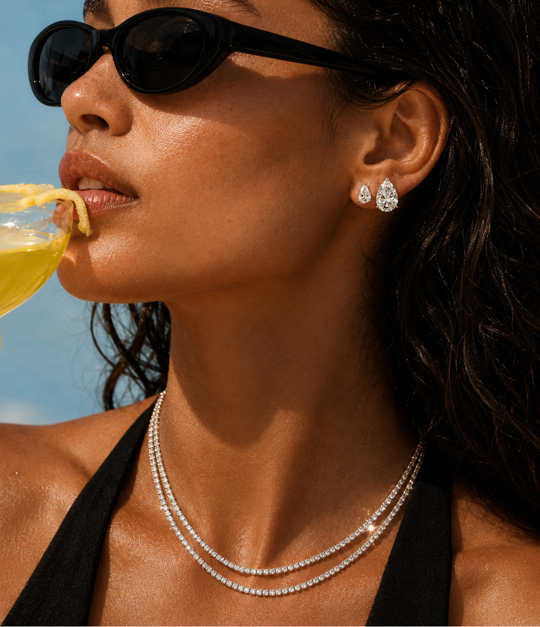 Woman wearing sunglasses and a necklace, drinking from a yellow cup against a blue sky and diamond tennis necklaces and a diamond pear cut stud earrings for euro summer